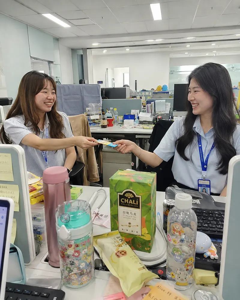 Two young office workers sharing a tea sachet during a workday break.